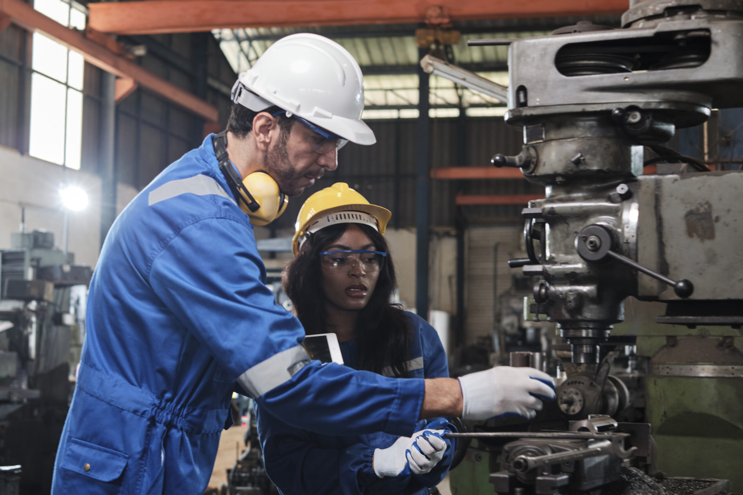 Two industrial workers in protective and safety uniforms and hardhats, male manager, and Black colleague work with metalwork machines in manufacturing factory. Professional production engineer team.